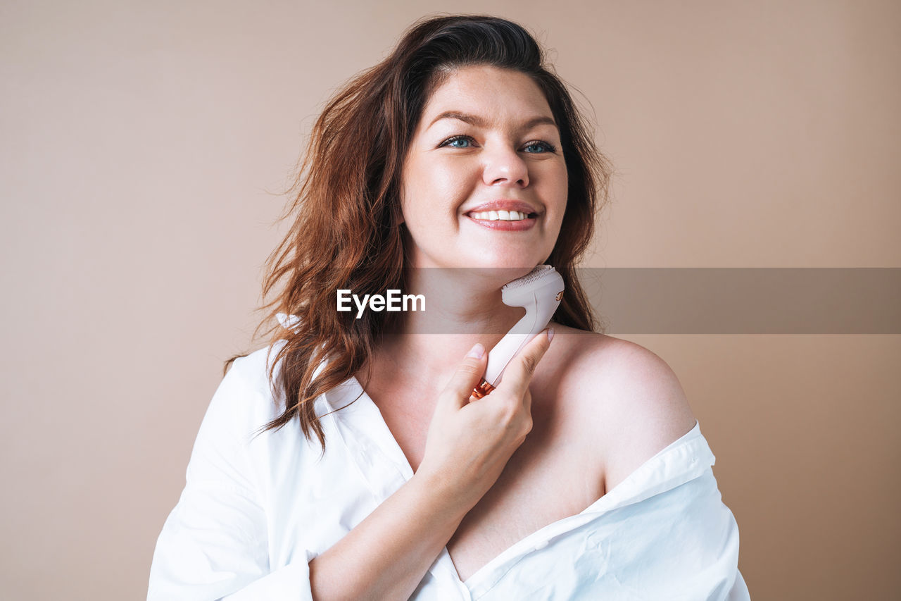 Self loving woman plus size in white shirt with facial massager in hands on beige background