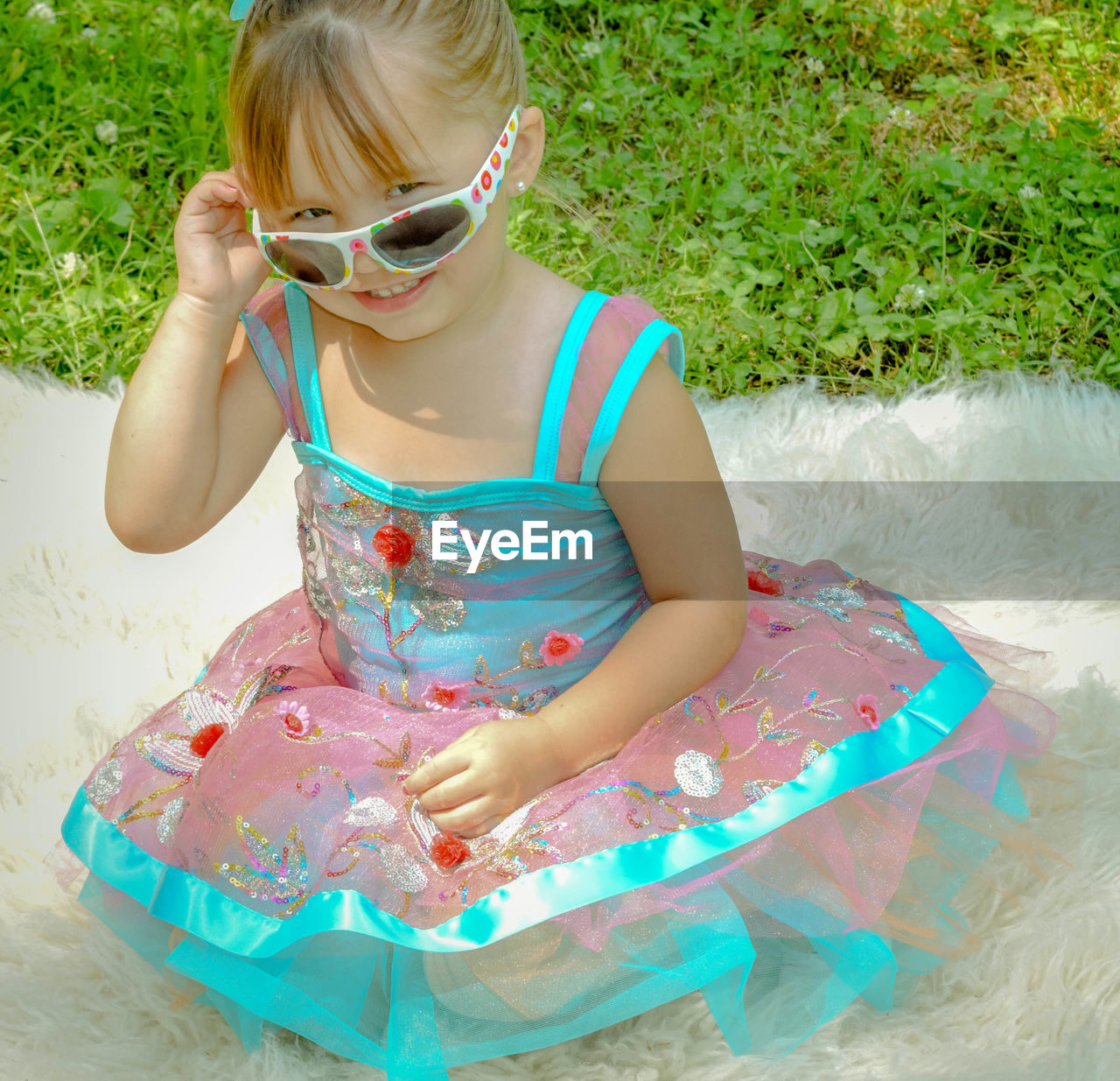 Full length portrait of cute smiling girl wearing sunglasses while sitting on rug at field