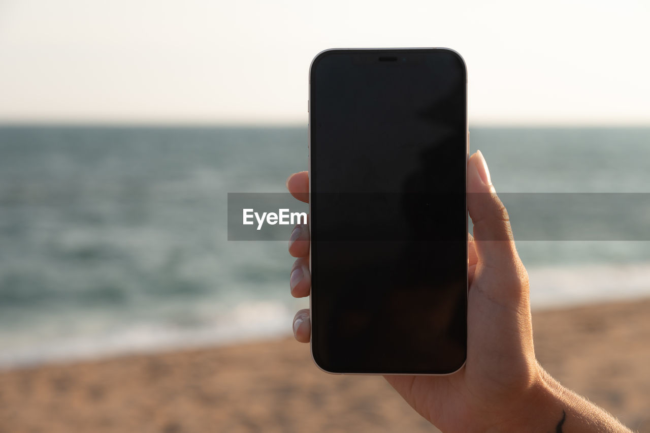 A girl holds a smartphone in her hand close-up with a black screen against the background of the sea