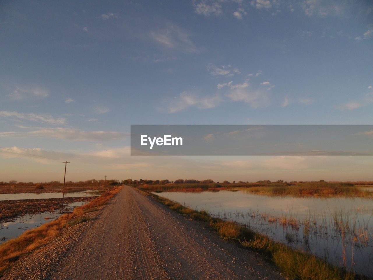 Empty dirt road by lake against sky