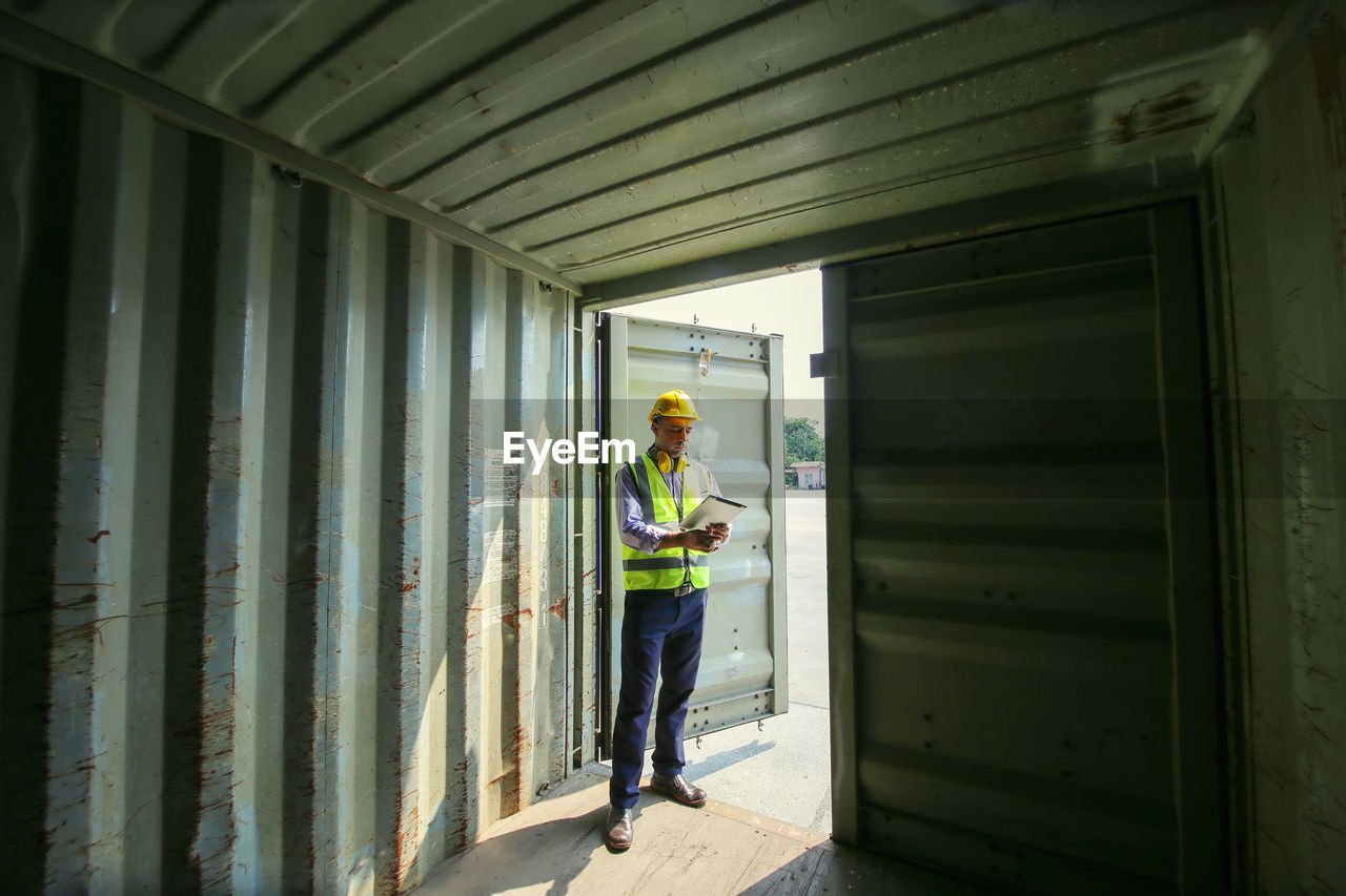 Full length of manual worker holding clipboard standing under cargo container