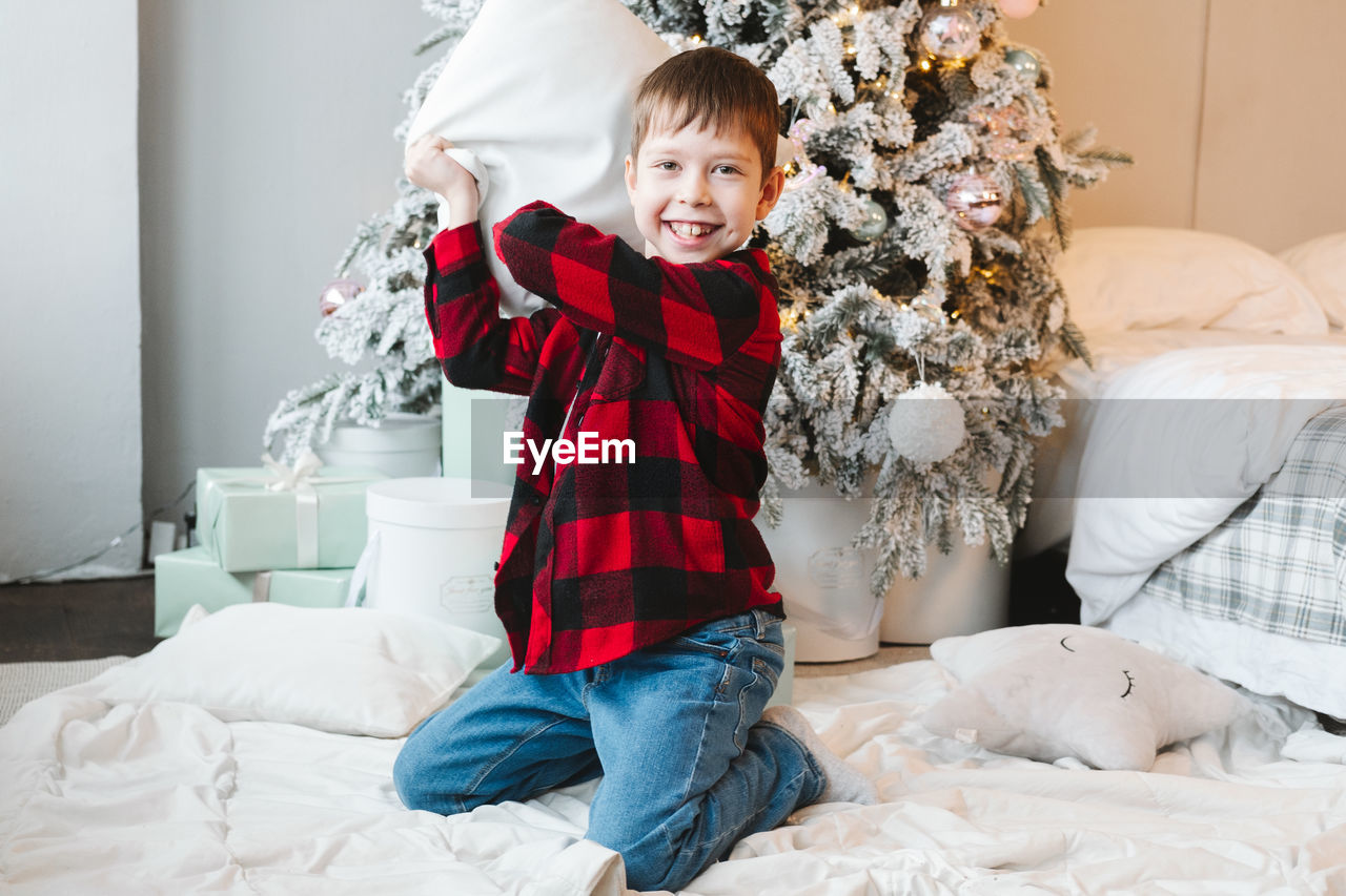 Boy in a red  shirt sits by the christmas tree with gifts on the floor and fights with a pillow