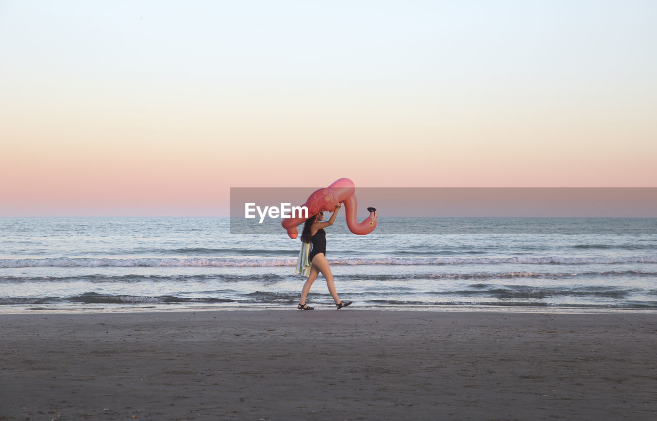 Young woman walking with inflatable pink flamingo on the beach at sunset