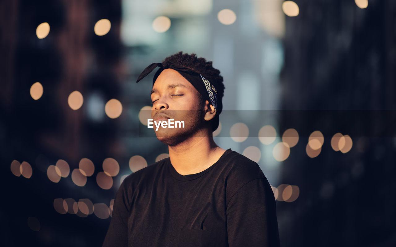 Young man with eyes closed standing against illuminated lights at night