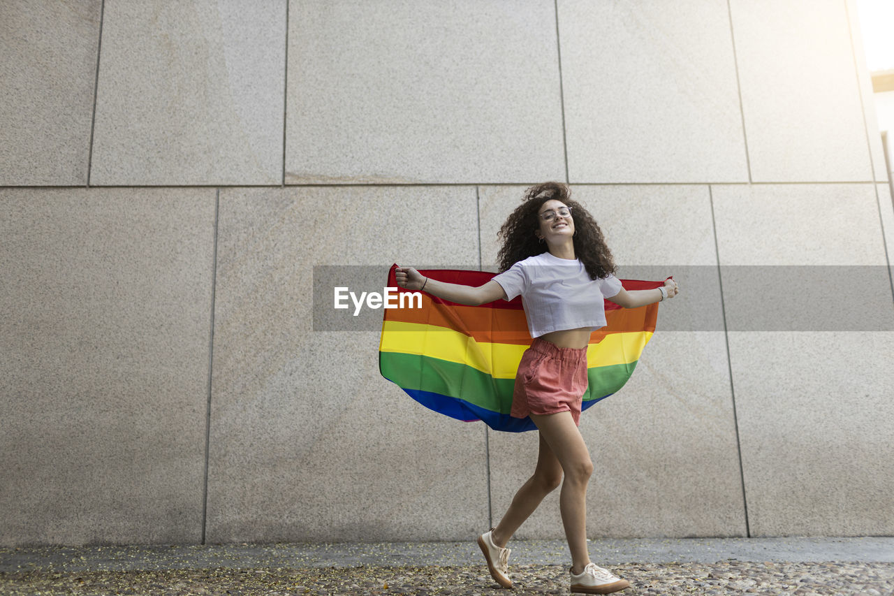 Woman holding rainbow flag while walking in front of wall