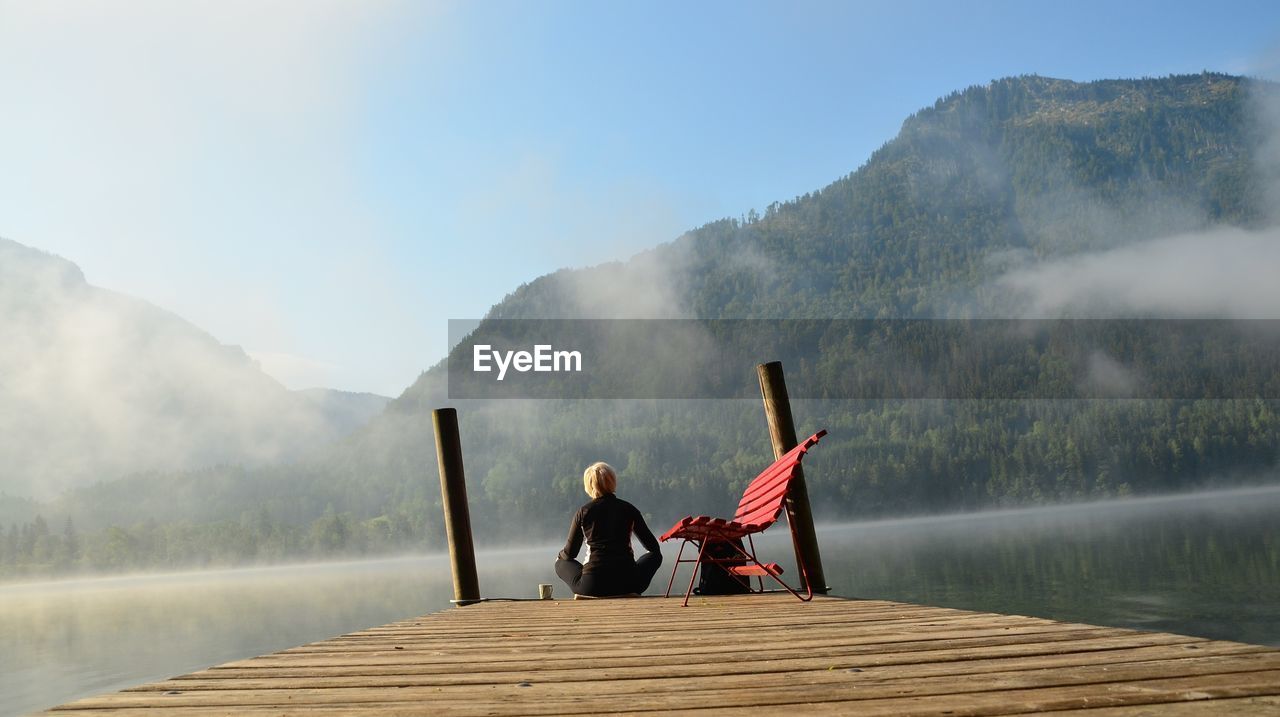 Rear view of woman sitting on pier at lake against mountain