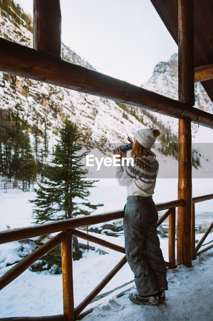 Female traveler enjoying hot drink from thermos while resting in wooden shelter near snowy mountains in banff national park