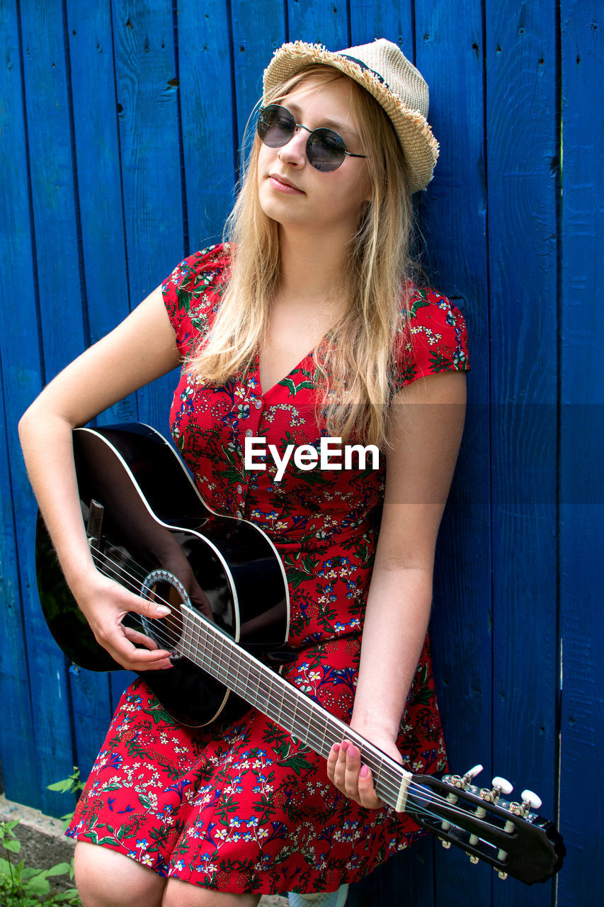 Beautiful teenage blonde long haired girl playing acoustic guitar outdoor over blue wooden fence. 