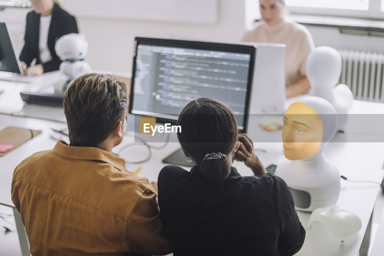 Rear view of multiracial male and female students discussing over computer in innovation lab