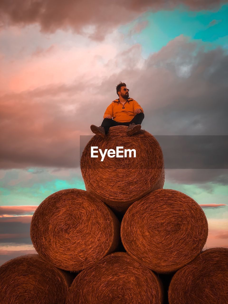 Man sitting on hay bales against sky during sunset