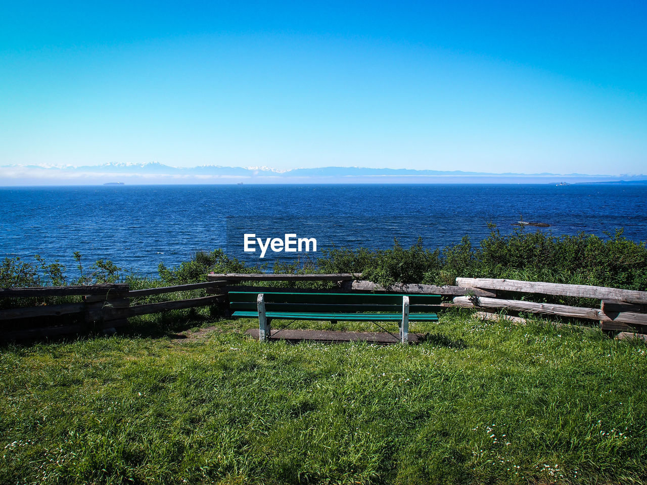 Bench on grassy field by sea against clear blue sky