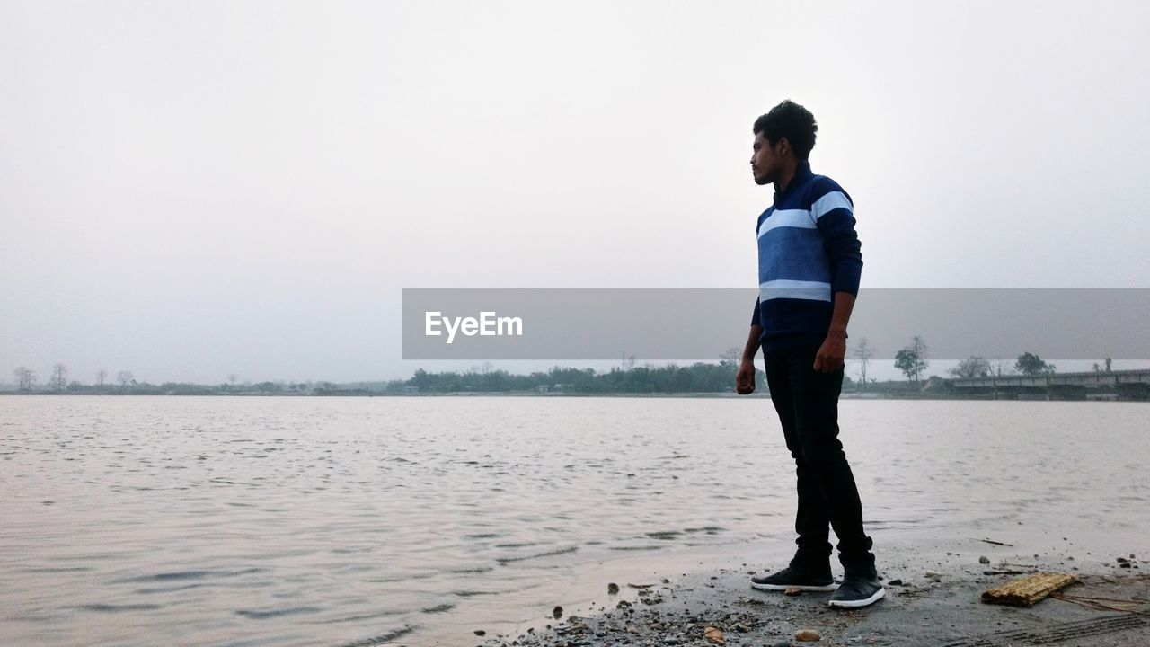 YOUNG MAN STANDING ON SHORE AGAINST CLEAR SKY