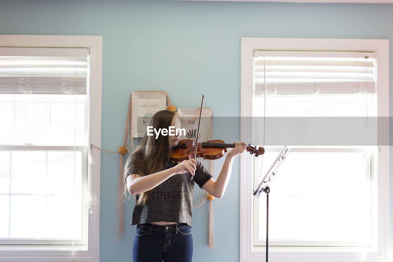 Woman playing violin while standing by window at home