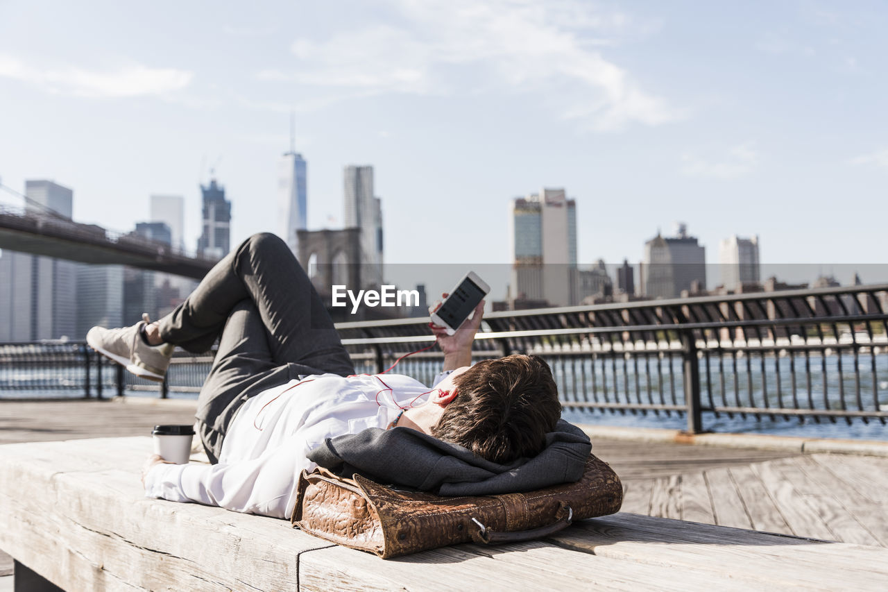 Usa, brooklyn, businesswoman lying on bench listening music with earphones