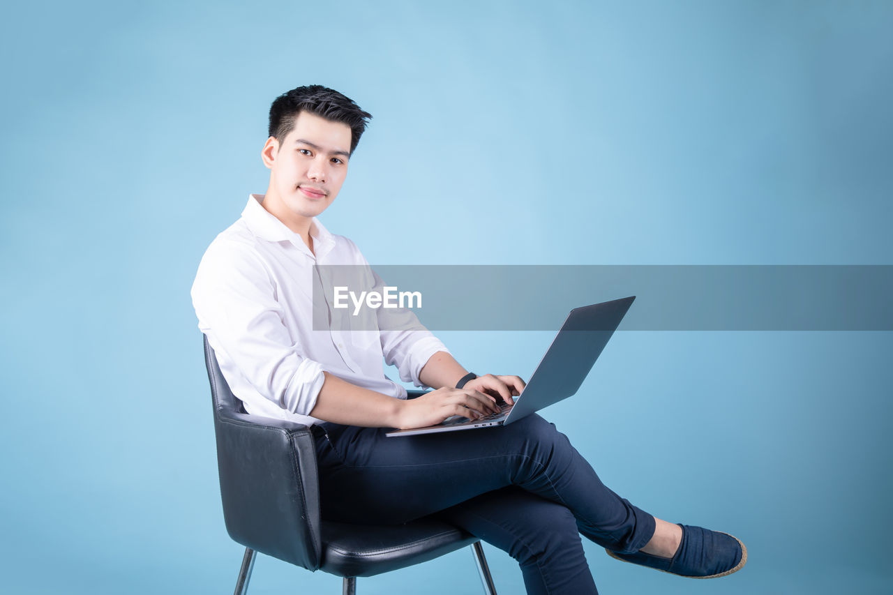 Portrait of young man using laptop while sitting on seat against blue background
