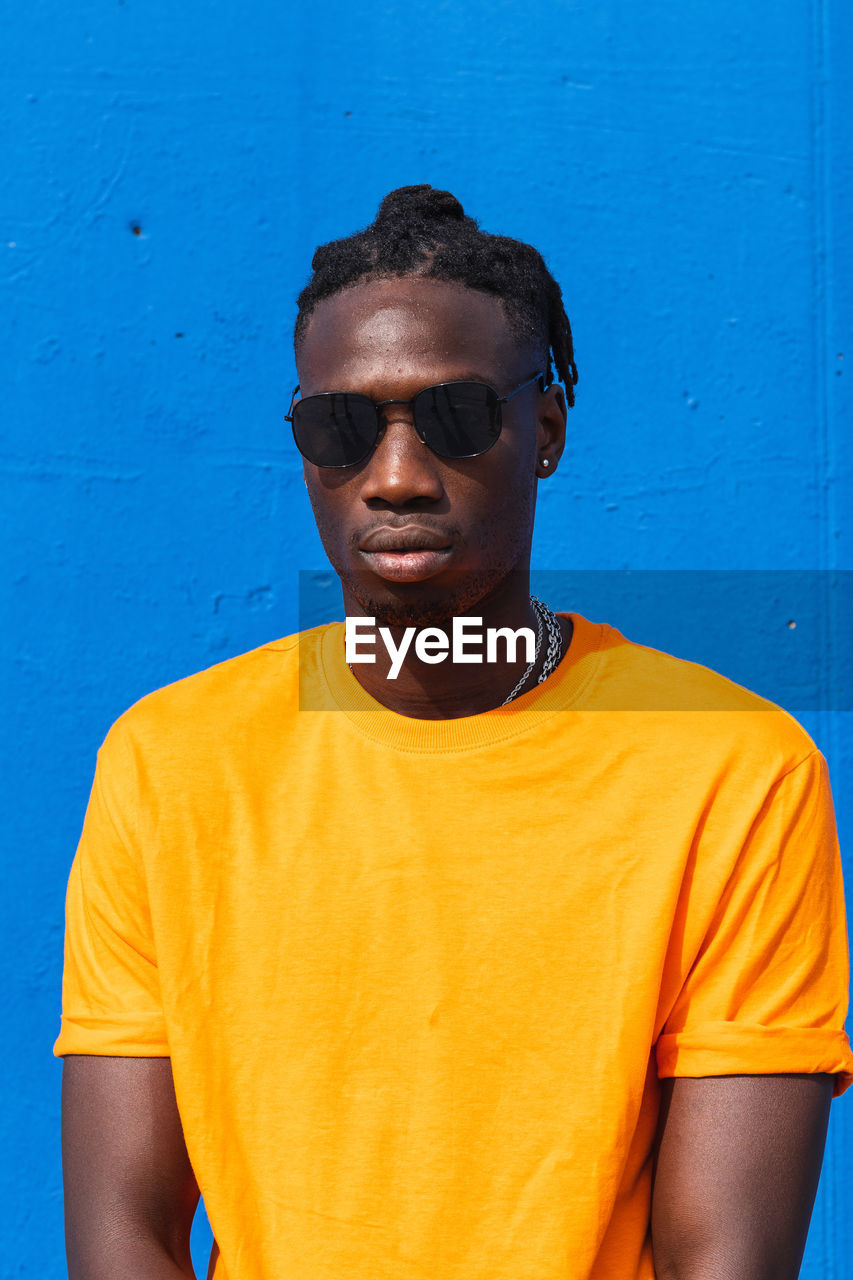 Serious young african american guy in bright yellow t shirt and trendy sunglasses looking at camera against blue background