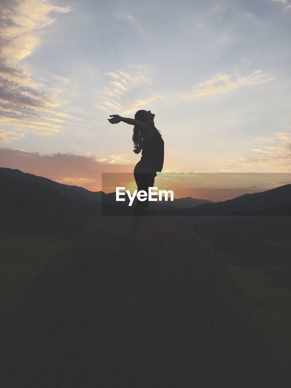 Side view of woman with arms outstretched standing on heap by mountains during sunset