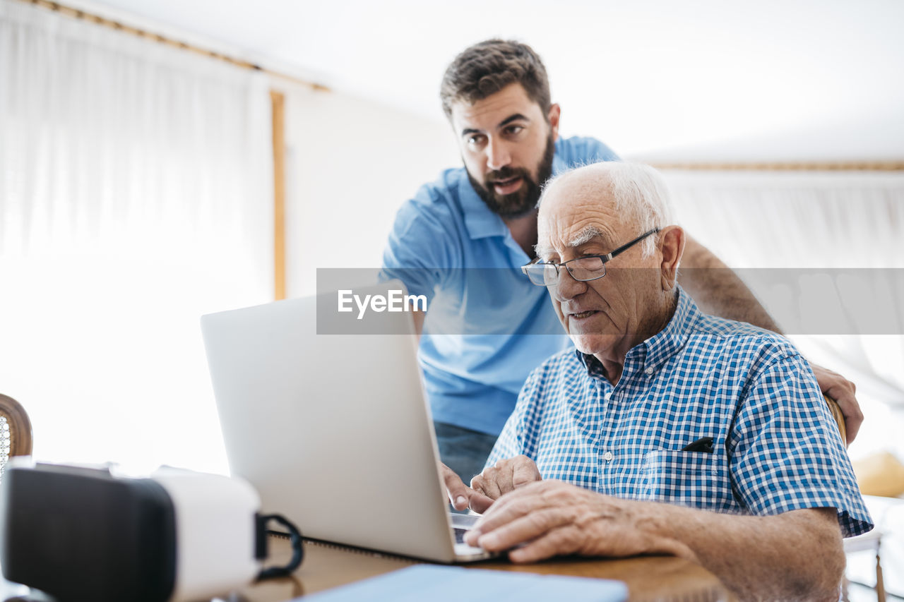 Adult grandson teaching his grandfather to use laptop