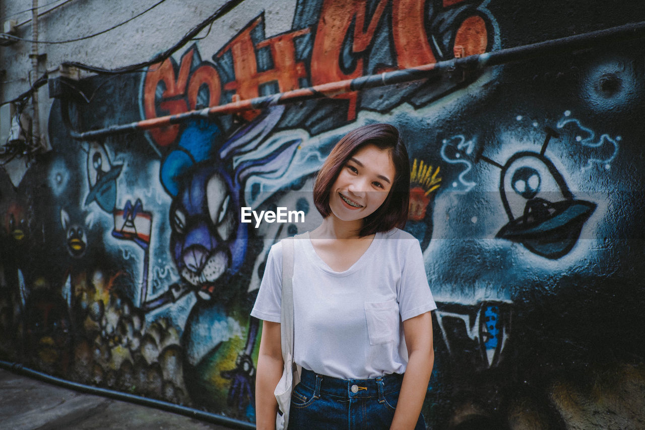 PORTRAIT OF YOUNG WOMAN STANDING AGAINST GRAFFITI WALL