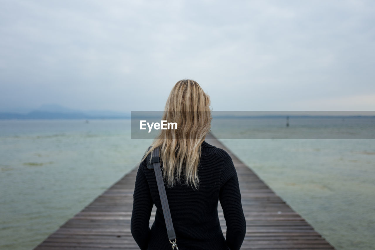 Rear view of mid adult woman standing on pier over sea against cloudy sky