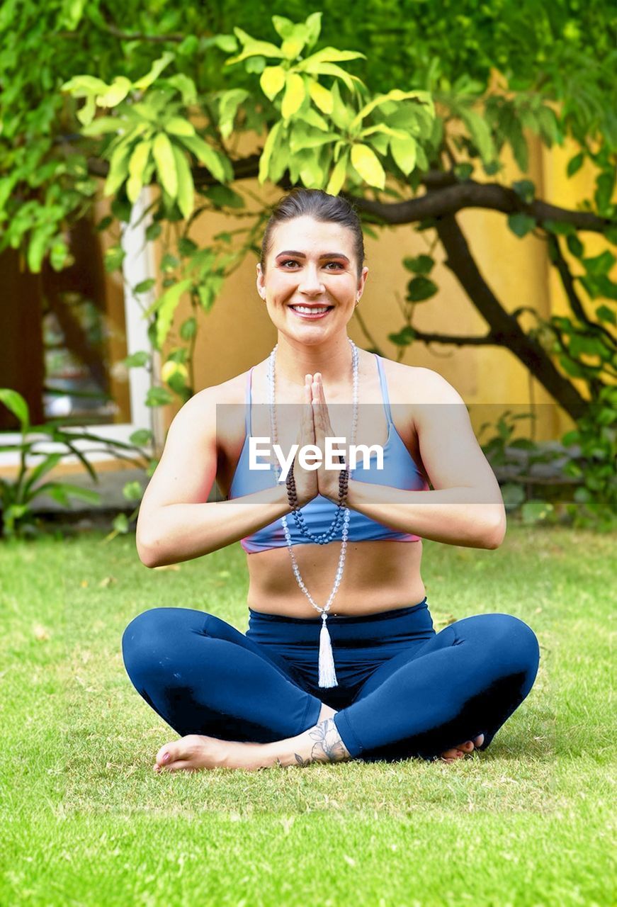 exercising, yoga, wellbeing, one person, adult, lifestyles, women, sports clothing, sports, sitting, zen-like, plant, relaxation, meditating, physical fitness, clothing, nature, grass, flexibility, spirituality, posture, smiling, full length, cross-legged, leisure activity, stretching, portrait, female, happiness, young adult, vitality, exercise mat, practicing, tree, lotus position, sports training, outdoors, religion, balance, green, front view, park, tranquility, person, day, martial arts, summer, park - man made space, emotion, looking at camera, harmony, athlete, tranquil scene, strength, hairstyle, healthcare and medicine, blond hair, cheerful, body care