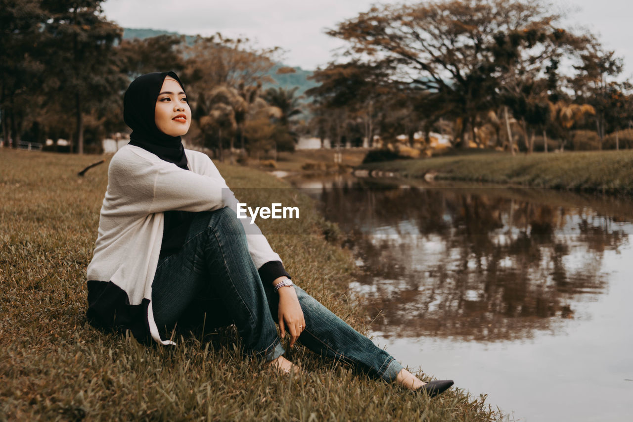 Young woman looking away while sitting by lake against trees