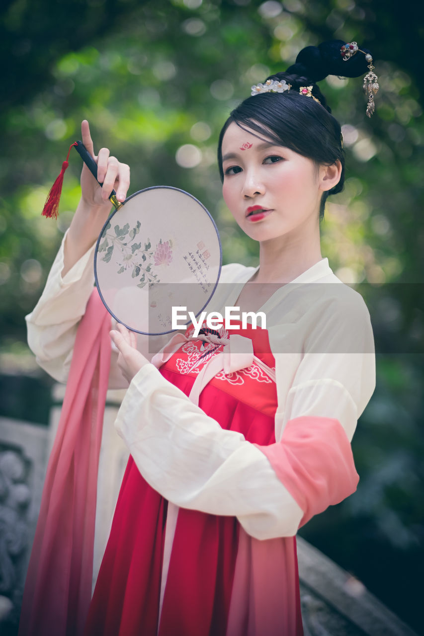 Portrait of woman in traditional clothing holding hand fan against tree