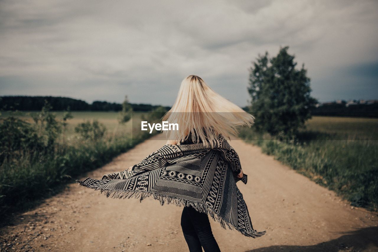 Young blond woman on dirt road against sky