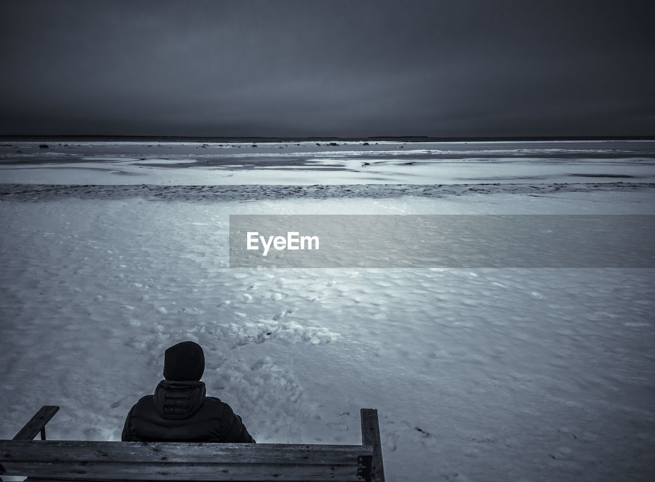 Rear view of boy sitting on bench against snow covered beach