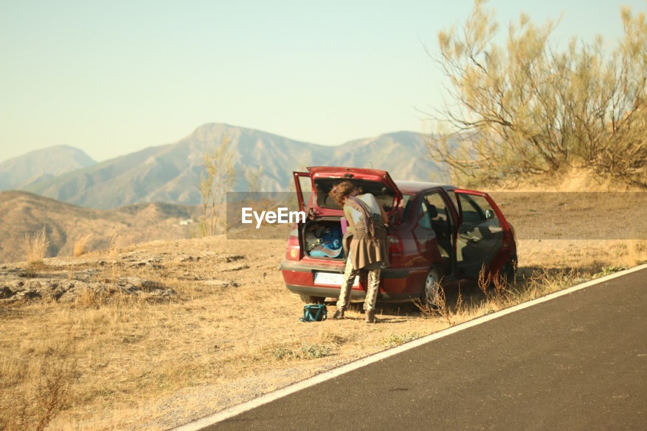 Rear view of woman standing by car on roadside with mountains in background