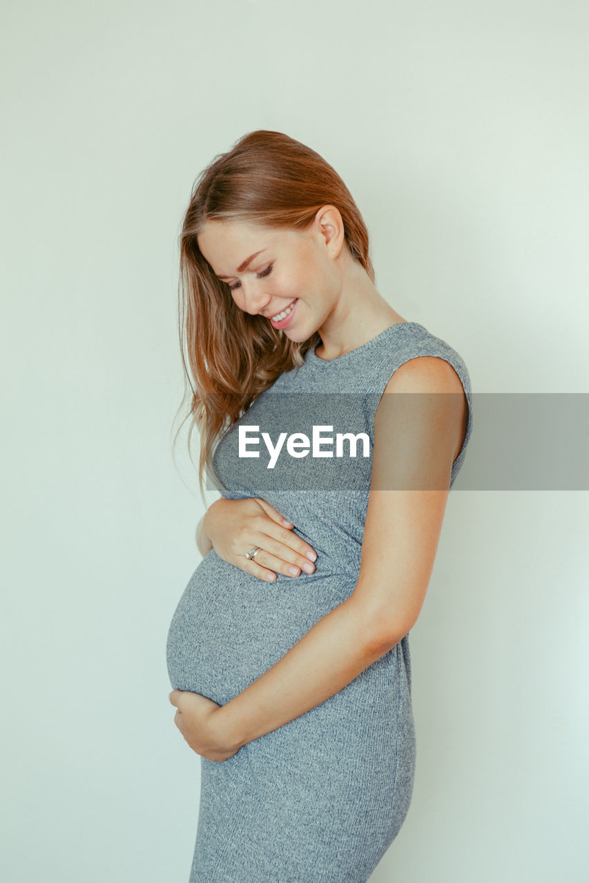 Pregnant young woman touching abdomen while standing against white background