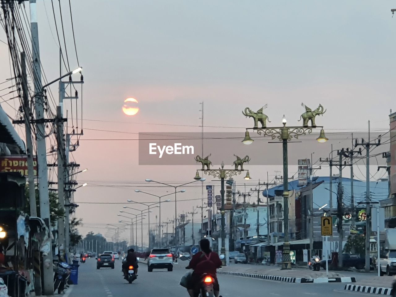 transportation, city, mode of transportation, sky, architecture, street, cityscape, car, motor vehicle, road, nature, infrastructure, built structure, building exterior, land vehicle, lighting, vehicle, street light, urban area, electricity, city street, evening, traffic, city life, cable, sunset, outdoors, downtown, travel, dusk, motion, light, sign, technology, electricity pylon, power line, lighting equipment, group of people, travel destinations, on the move, power generation