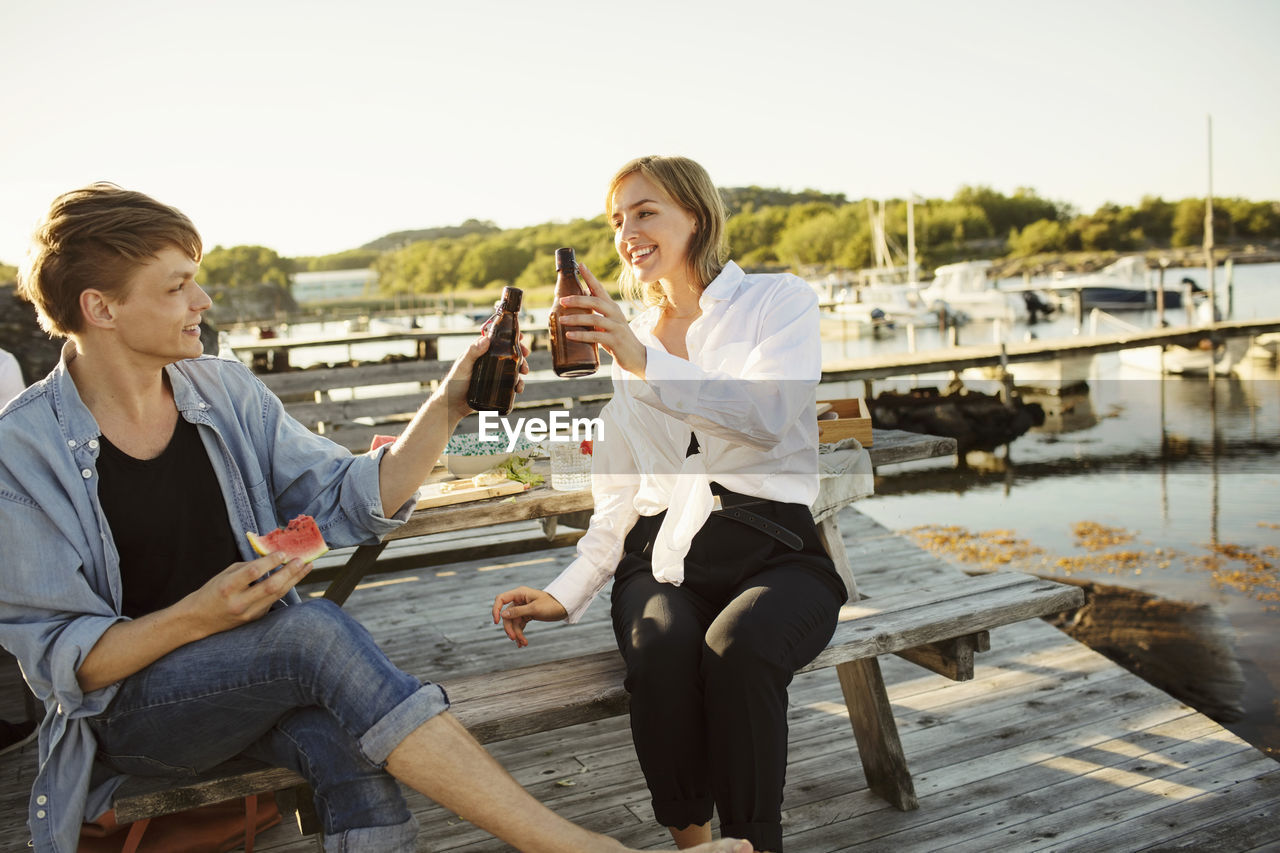 Smiling male and female friends toasting beer bottles at harbor