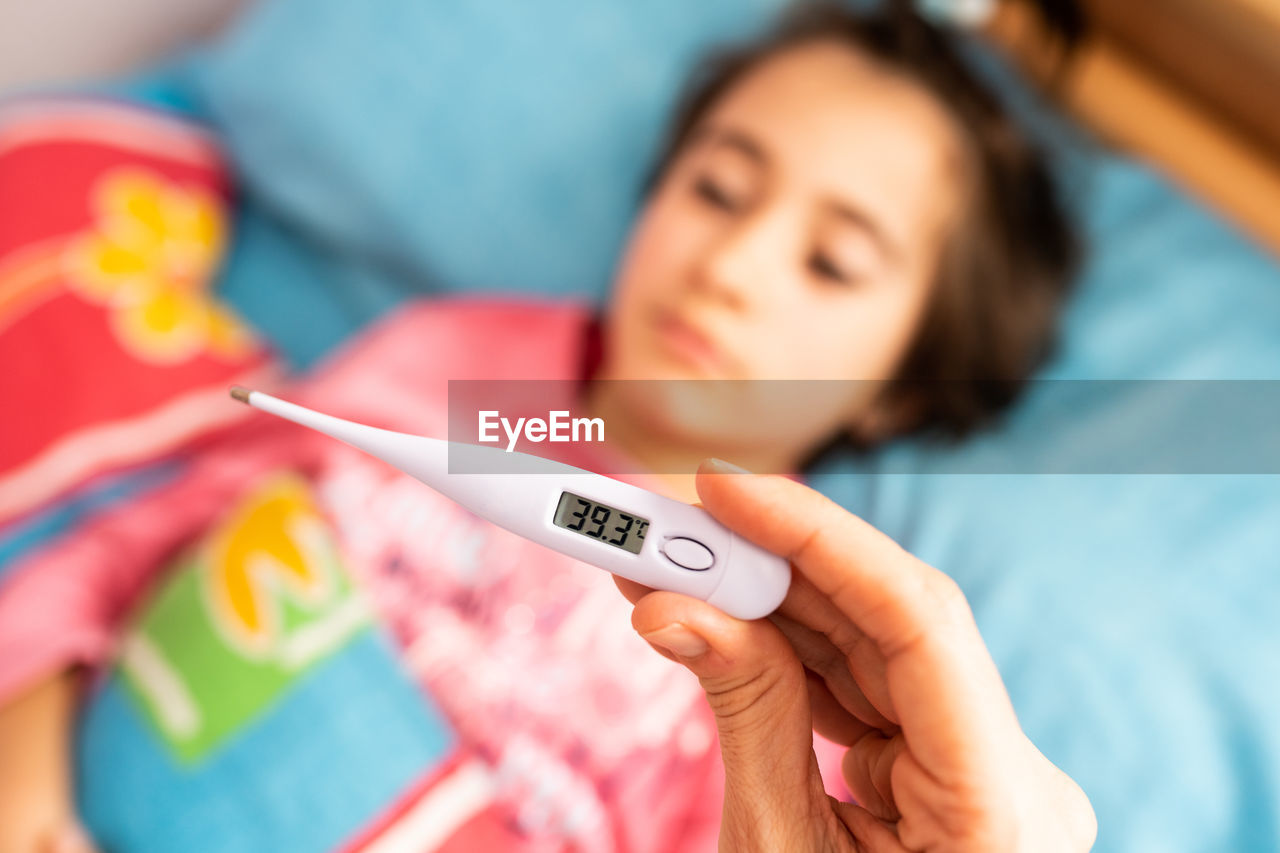 Close-up of woman hand holding thermometer over daughter lying on bed