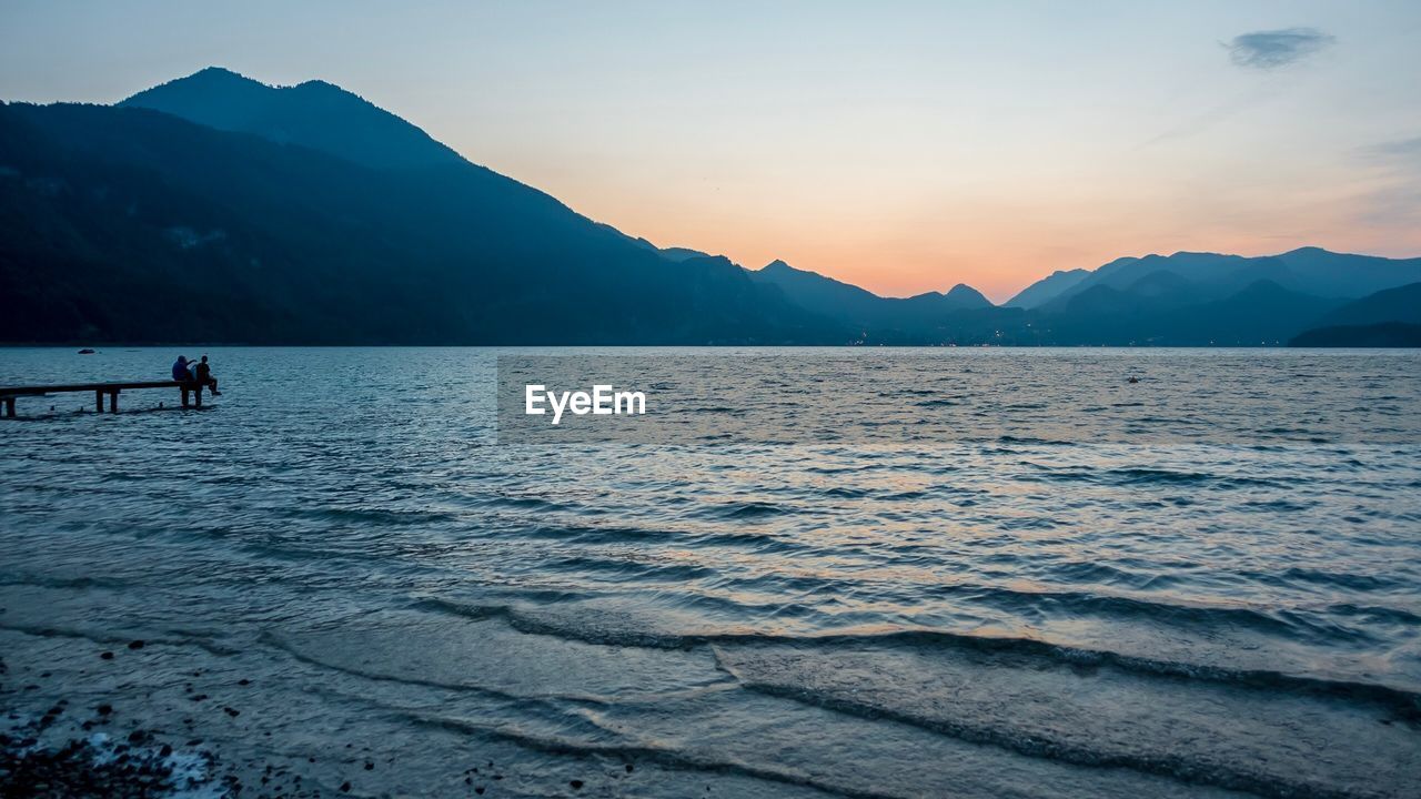 People sitting on pier by lake against sky during sunset