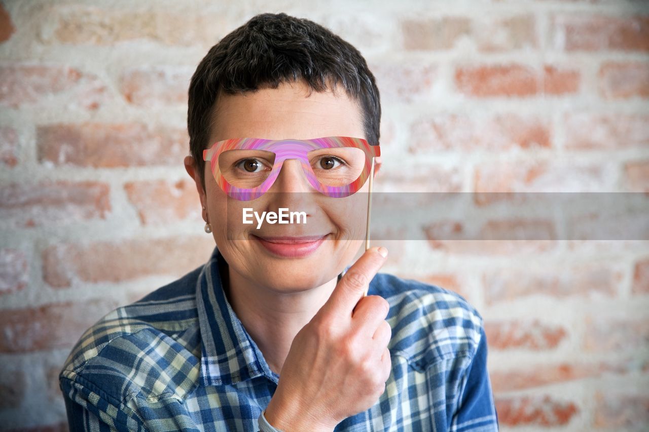 Portrait of smiling woman holding prop eyeglasses against wall