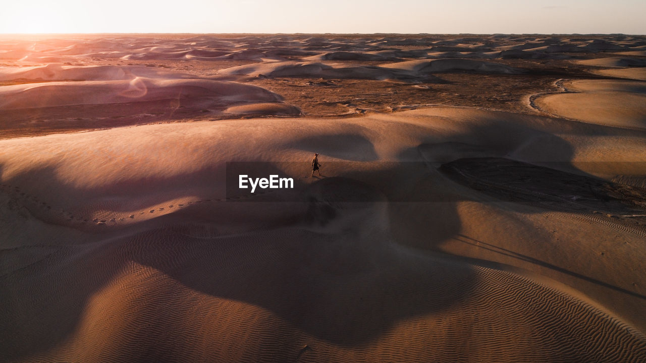 Aerial view of desert dunes with a man walking alone across a vast desert at sunset