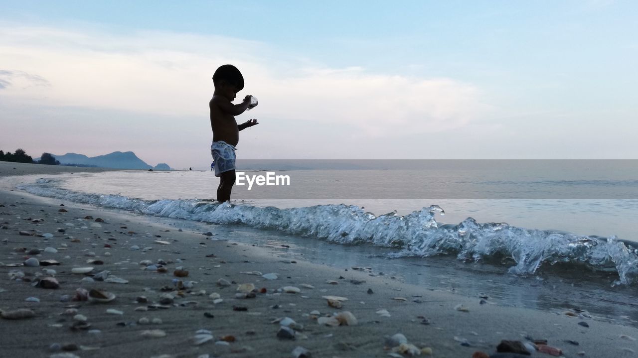Side view of boy standing on shore at beach against sky during sunset