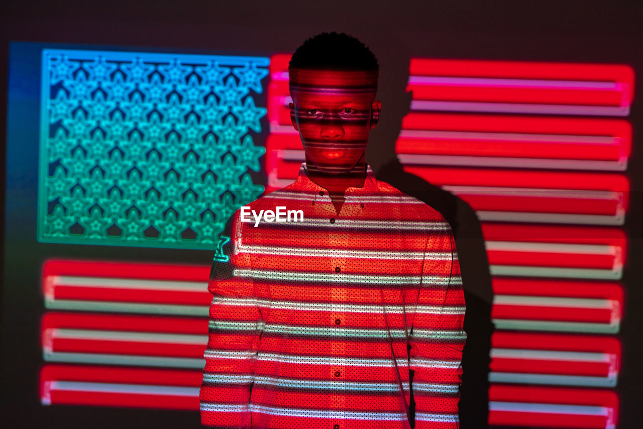 Serious african american male illuminated by red and blue neon projection of national american flag standing in dark studio