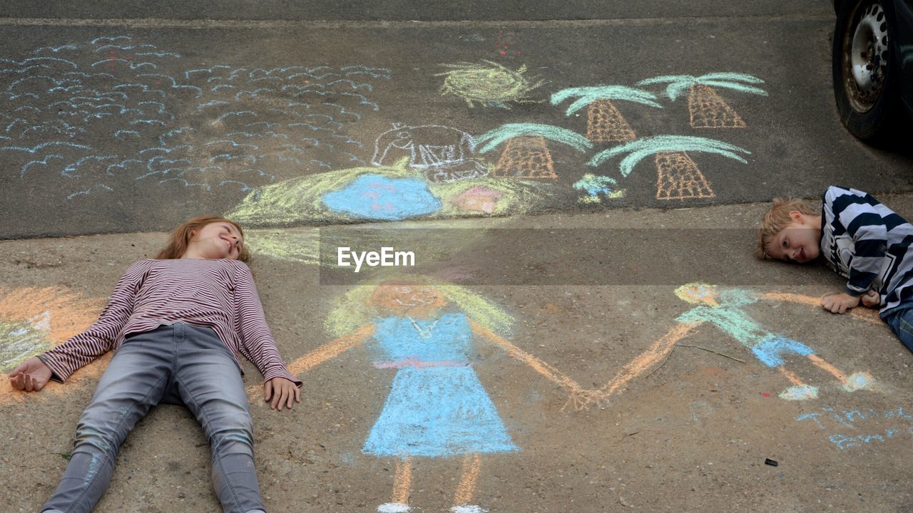 High angle view of siblings lying on chalk drawings at footpath