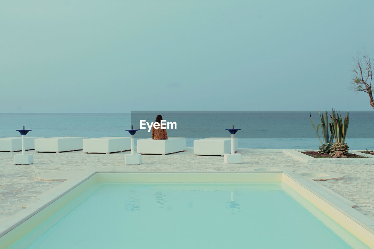 Rear view of woman sitting at poolside by sea against clear sky