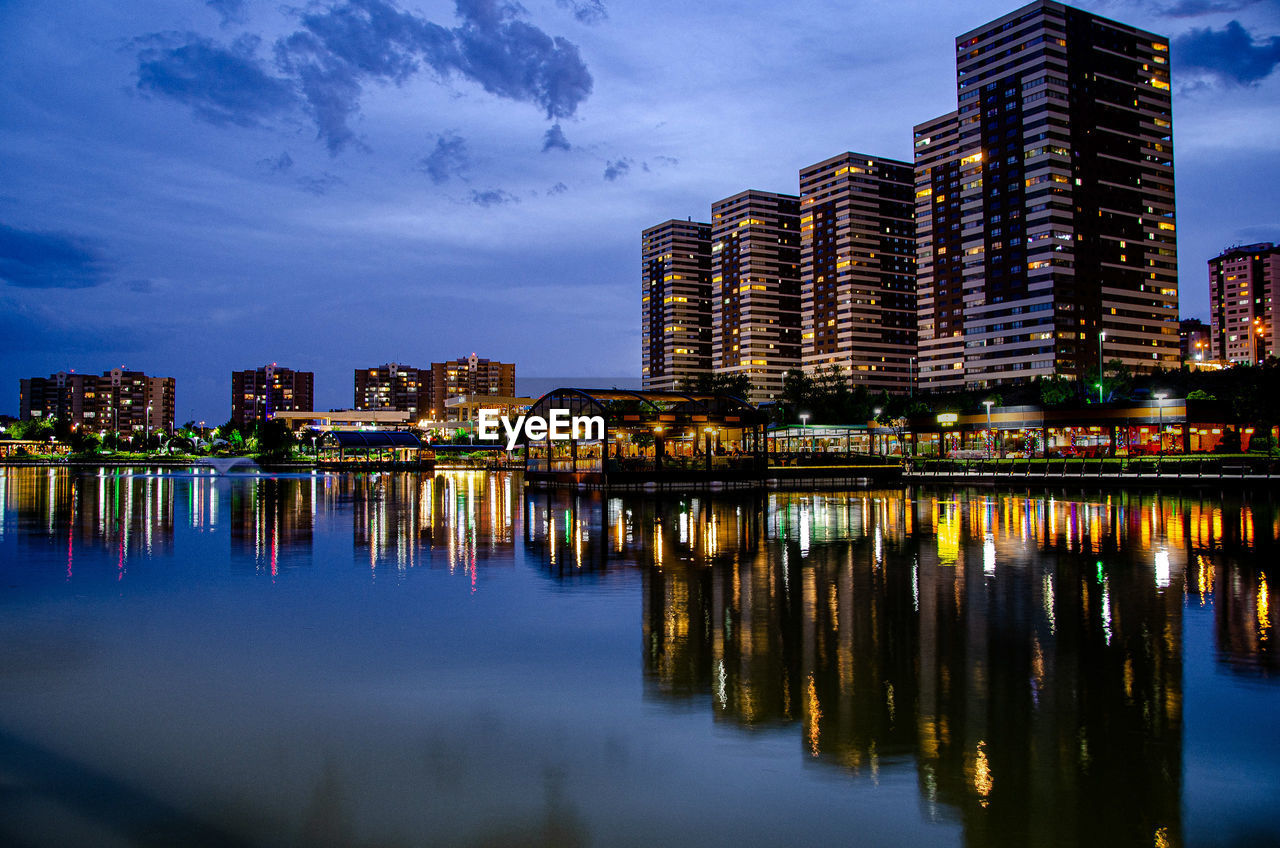 ILLUMINATED BUILDINGS BY RIVER AGAINST SKY