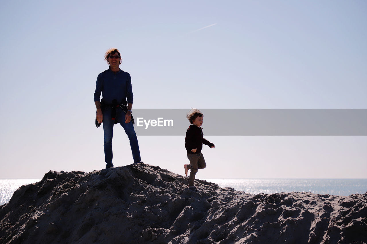 Father and son on rock formation at beach against clear sky
