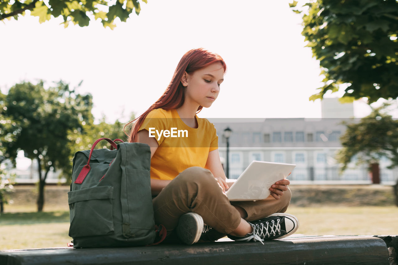 A teenage girl is sitting on a park bench with a laptop and preparing for lessons or exams.