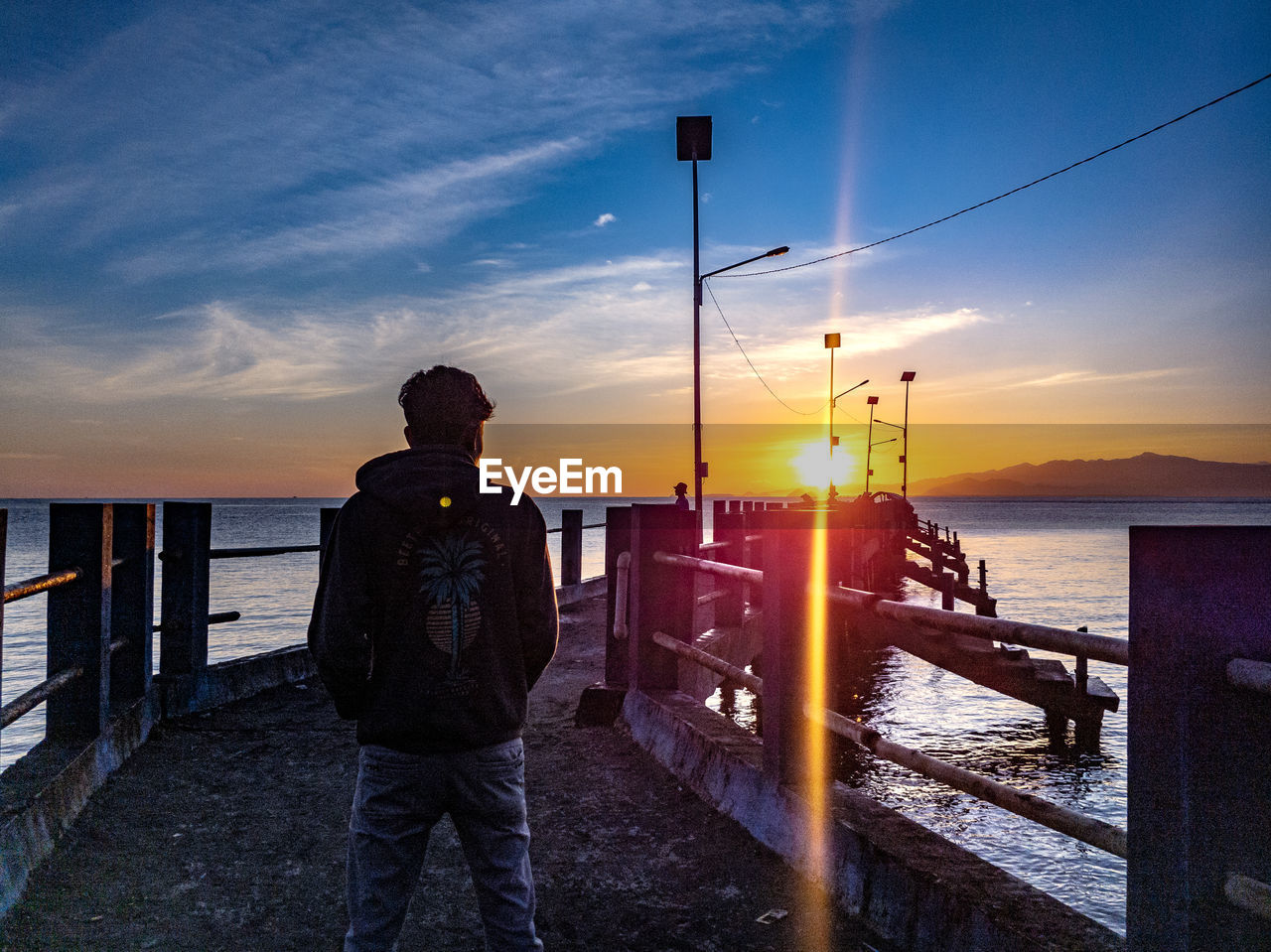 REAR VIEW OF MAN STANDING AT BEACH AGAINST SKY DURING SUNSET