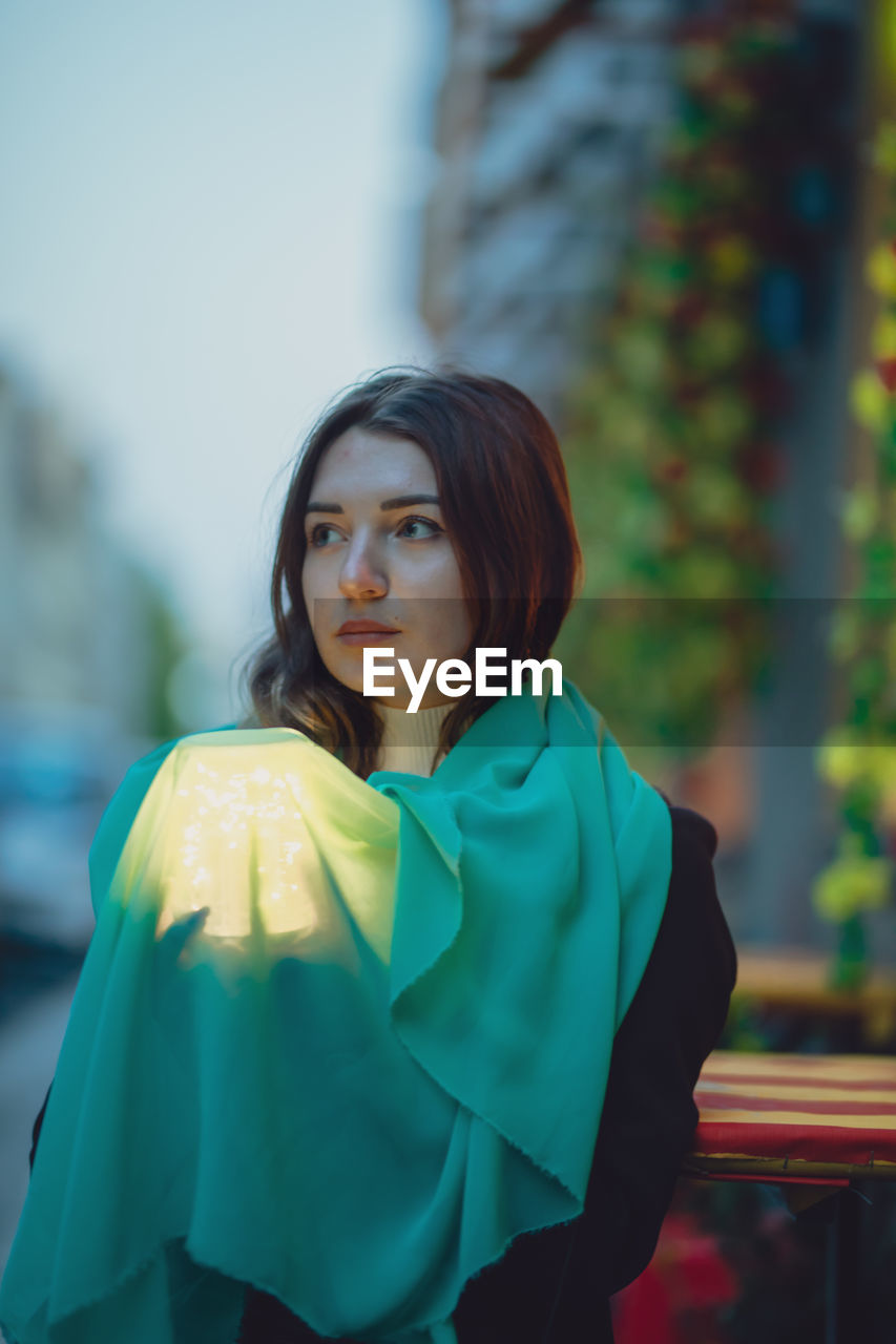 Thoughtful beautiful woman holding glowing jar under blue scarf in city at dusk