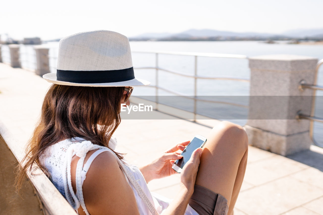 REAR VIEW OF WOMAN USING MOBILE PHONE WHILE SITTING ON DECK