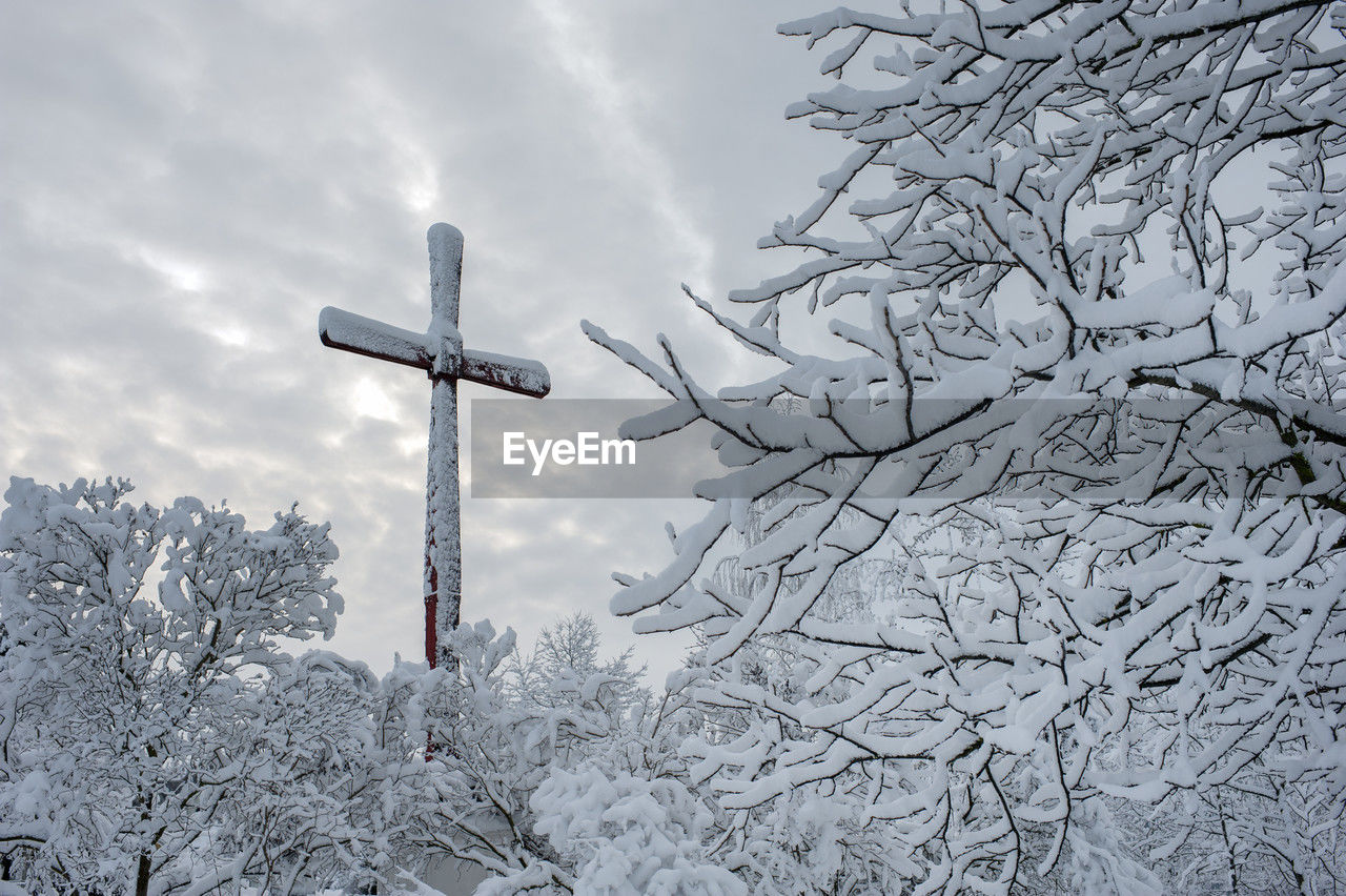 Wooden frozen cross completely covered with snow and icicles and winter landscape under bright sky.