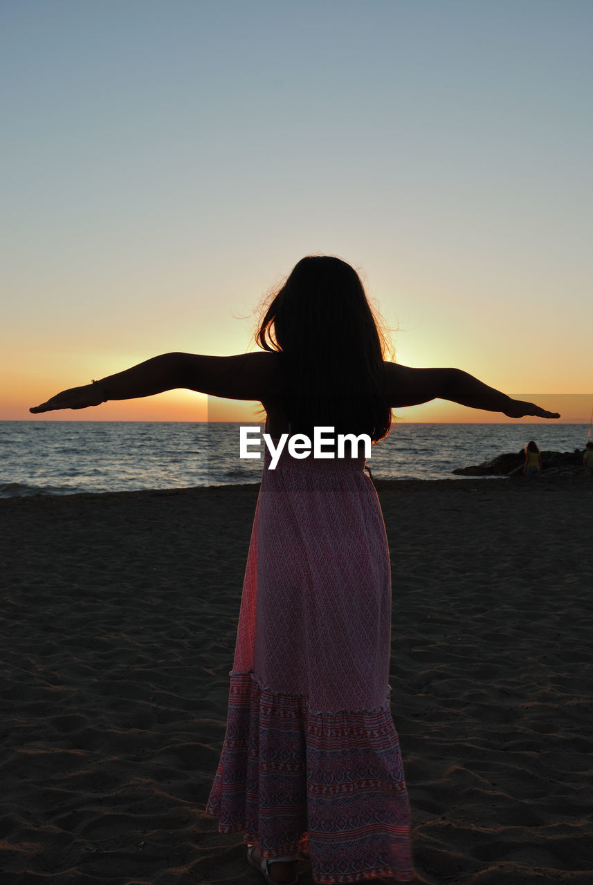 REAR VIEW OF WOMAN STANDING IN SEA AT SUNSET