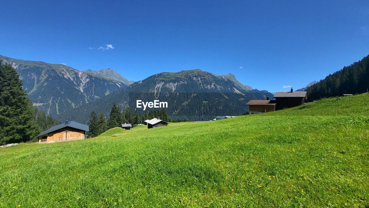 mountain range, mountain, scenics - nature, meadow, landscape, environment, plant, land, sky, grass, building, architecture, house, beauty in nature, nature, built structure, pasture, grassland, building exterior, field, green, valley, rural scene, plateau, hut, residential district, blue, no people, plain, highland, travel, travel destinations, tree, tranquility, village, ridge, tranquil scene, wilderness, summer, agriculture, non-urban scene, outdoors, mountain peak, tourism, environmental conservation, idyllic, farm, clear sky, forest, social issues, agricultural building, snow, rural area, sunlight, pine tree, cloud, mansion, cottage, mountain pass, activity, day